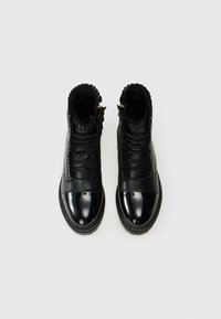 Botas de tobillo de cuero negro con una punta brillante, detalle de volantes texturizados alrededor del collar y cordones en la parte delantera. Cremalleras doradas a los lados.
