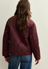 Burgundyrutig quiltad jacka med ett vågigt mönster, med en avslappnad passform och vinklade ärmar, bärs över blå denimjeans.