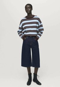 Donna in piedi con un maglione a maniche lunghe a righe blu e marroni, pantaloni cropped larghi e scuri, e stivaletti neri con lacci.