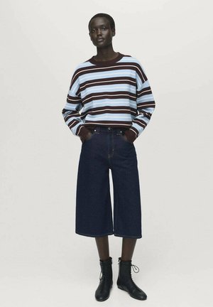 Vrouw die staat en een blauw en bruin gestreept langaf sweater, donkere wijde cropped broek en zwarte veterschoenen tot de enkel draagt.