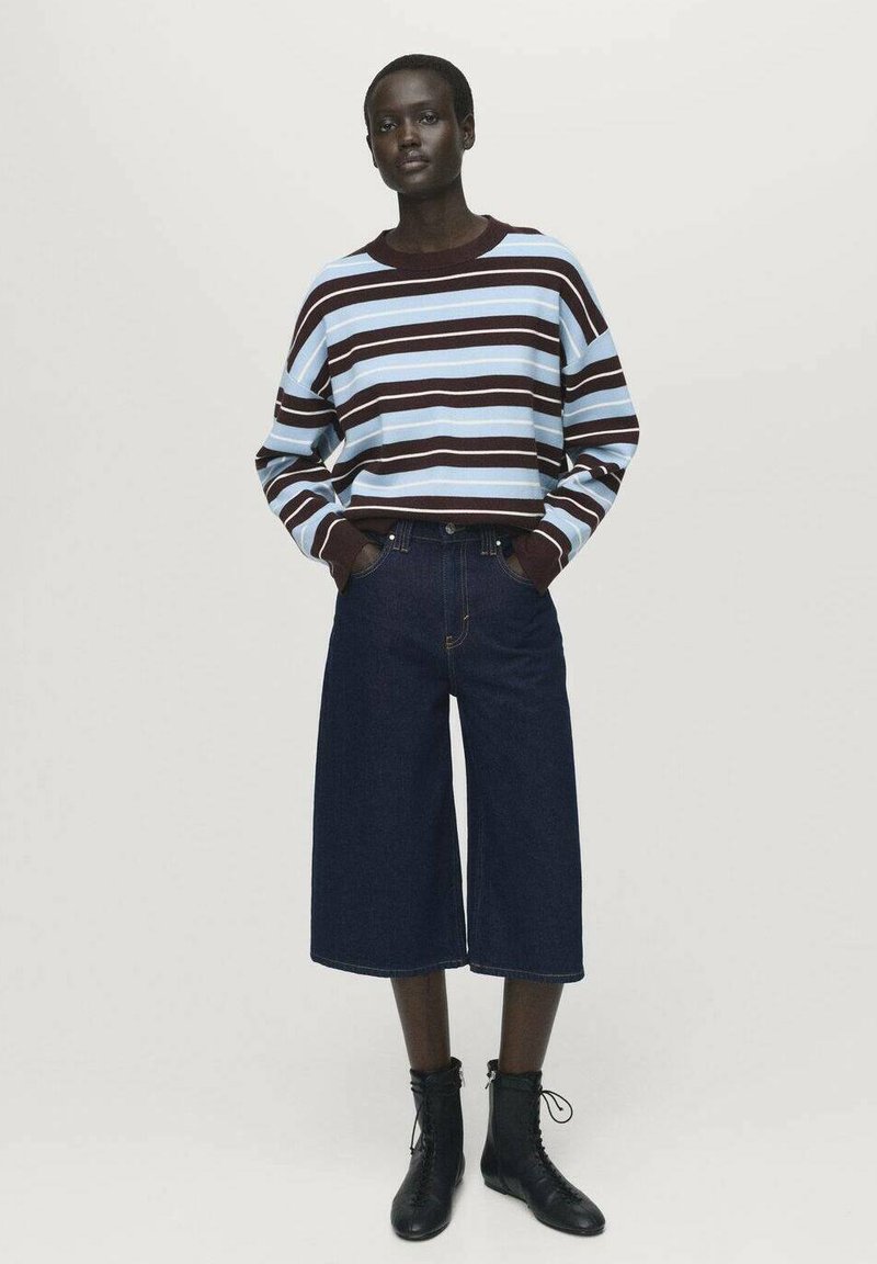 Donna in piedi con un maglione a maniche lunghe a righe blu e marroni, pantaloni cropped larghi e scuri, e stivaletti neri con lacci.