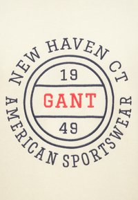 Κρεμ υφασμα με κυκλικό έμβλημα που περιλαμβάνει τις λέξεις "NEW HAVEN CT", "GANT" με κόκκινα γράμματα, τους αριθμούς "19" και "49", και τη φράση "AMERICAN SPORTSWEAR" με μπλε σκούρα γράμματα.