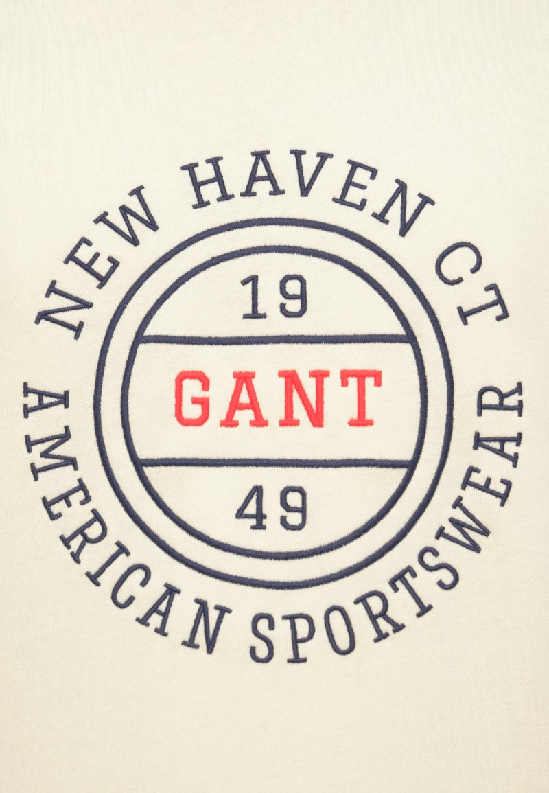 Tissu couleur crème présentant un emblème circulaire avec les inscriptions "NEW HAVEN CT", "GANT" en rouge, "19" et "49", ainsi que "AMERICAN SPORTSWEAR" en texte bleu marine.