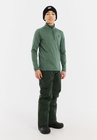 Maglietta verde a maniche lunghe con zip abbinata a pantaloni verdi scuri. Il materiale sembra liscio, design aderente e completato da un berretto nero.