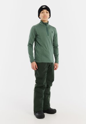 Maglietta verde a maniche lunghe con zip abbinata a pantaloni verdi scuri. Il materiale sembra liscio, design aderente e completato da un berretto nero.