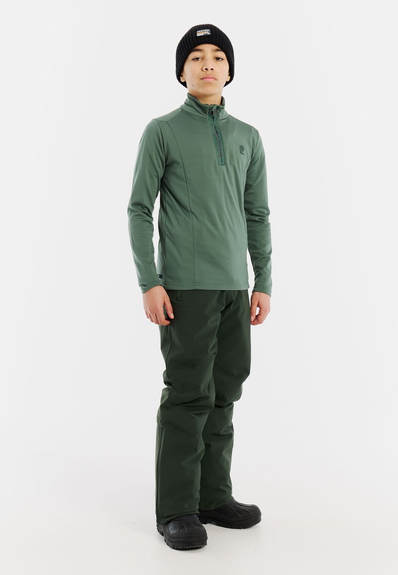 Maglietta verde a maniche lunghe con zip abbinata a pantaloni verdi scuri. Il materiale sembra liscio, design aderente e completato da un berretto nero.