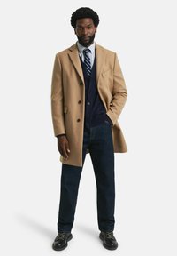 Beiger Wollmantel über einer marineblauen Weste und einem gestreiften Hemd, kombiniert mit dunkler Jeans und schwarzen Schnürschuhen. Mann steht selbstbewusst.