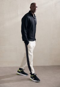 Czarna kurtka na zamek, białe spodnie z szarym bocznym paskiem oraz czarne sneakersy z neonowymi akcentami. Gładki materiał i sportowy krój.