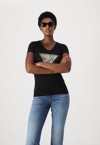 T-shirt nera a V con grafica testurizzata con logo "GUESS" e stampa leopardata. Il modello l'ha abbinata a jeans blu e occhiali da sole neri.