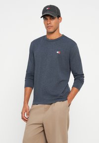 Jūras spēku zilas krāsas krekls ar garām piedurknēm, izgatavots no mīkstas, teksturētas auduma, ar mazu izšūtu logotipu sarkanā, baltā un zilā krāsā. Apvienots ar beigu krāsas brīva griezuma biksēm un atbilstošu cepuri.