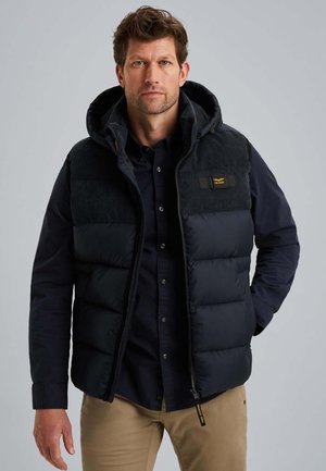 Homme portant un gilet matelassé noir sur une chemise à boutons sombre et un pantalon beige, se tenant devant un fond gris uni.