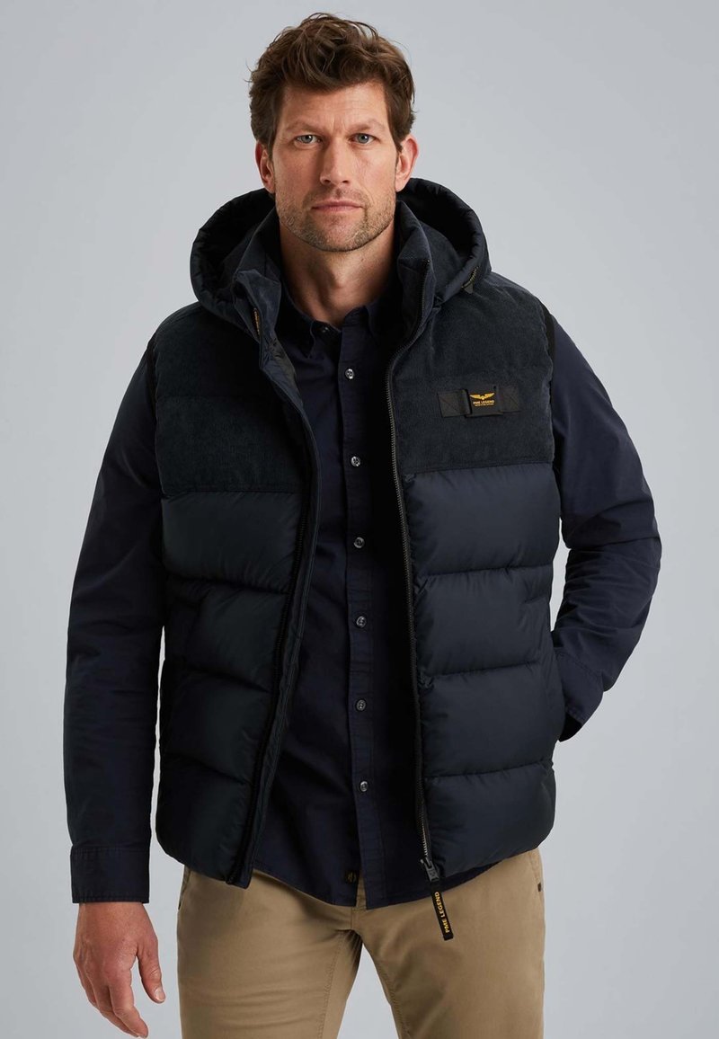 Man in een zwart gewatteerd vest over een donkere button-up shirt en tan kleurige broek, staande tegen een effen grijze achtergrond.