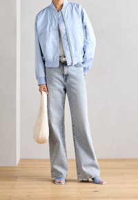 Jasnoniebieska kurtka bomber z prążkowanymi mankietami, szara bluzka, jasnoniebieskie jeansy o szerokich nogawkach, beżowa kopertówka oraz niebieskie sandały z odkrytymi palcami.