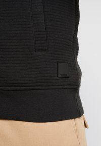 Sort pullover med ribbet tekstur, der har en lille sort label i nederste venstre hjørne, parret med lysebeige bukser.