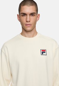 Sudadera color crema con cuello redondo y hombros caídos. Presenta un logo bordado de FILA en rojo, azul y blanco en el pecho.