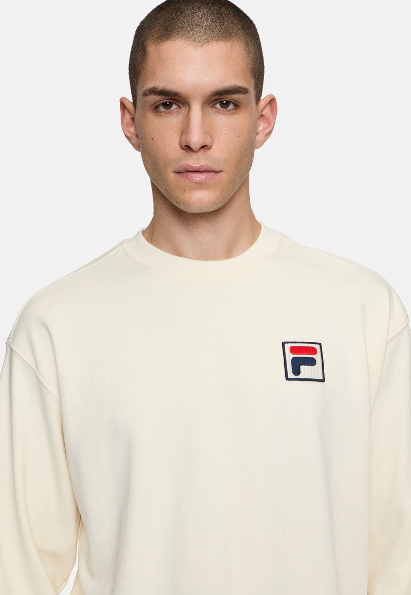 Felpa color crema con collo rotondo e spalle scese. Presenta un logo FILA ricamato in rosso, blu e bianco sul petto.
