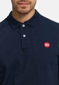 Tummansininen poolopaita, jossa on teksturoitu kangas, kaksinappinen aukko, valkoiset napit ja vasemmassa rinnassa pyöreä punainen kirjailtu logo.