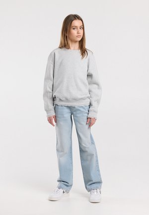 Jeune personne debout portant un sweat-shirt gris clair, un jean bleu clair et des baskets blanches sur un fond blanc uni.