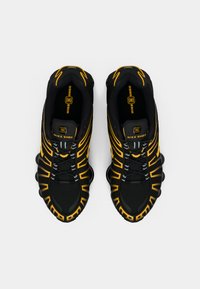 Melnas un dzeltenas Nike Shox kedas ar acs virsu, unikālu viļņu rakstu, mīkstu apkakli un apaļu mīkstinātu zoli.