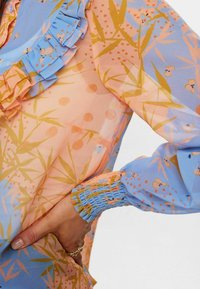 Långärmad genomskinlig blus med pastellblå och persikofärgade nyanser samt blommönster och bambumönster, rynkad krage och veckade ärmslut.