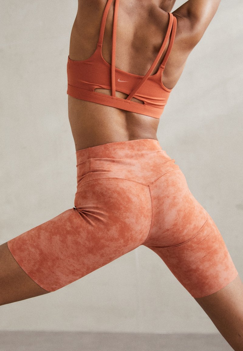 Personne athlétique s'étirant avec les bras levés, portant un soutien-gorge de sport orange à double bretelles et un short cycliste taille haute assorti.