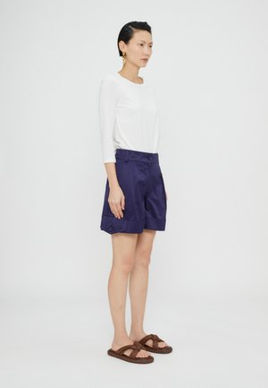 Mørkeblå shorts med folder og sidespenner, kombinert med en hvit langermet topp. Modellen har på seg brune sandaler, som viser en avslappet stil.