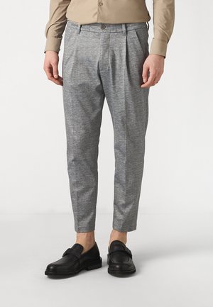 Mand iført skræddersyede grå bukser, beige skjorte og sorte læder loafers, stående på en lys baggrund.