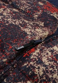 Doudoune avec une base bleu marine, texturée avec des motifs éclaboussés rouges et beiges. Dotée d'une fermeture éclair noire et d'une étiquette visible avec le logo "VANS".