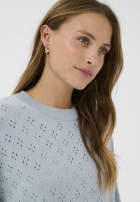 Pull en maille bleu clair avec des motifs ajourés, col rond et poignets côtelés, assorti de boucles d'oreilles en or.