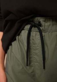 Shorts verts légers avec une taille élastique, un cordon noir et un détail subtil de texture. Le haut noir est partiellement visible au-dessus.