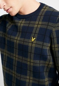 Pull en maille bleu marine avec un motif à carreaux vert et noir, arborant un petit logo brodé jaune sur la poitrine.