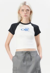 Vit kortärmad t-shirt med svarta korta ärmar, med blå texten "COSMIC DREAMS Obey" på framsidan, i kombination med grå byxor med snörning.