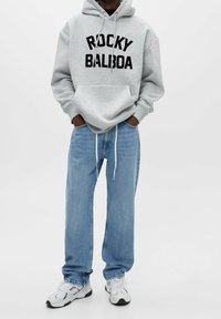 Personne portant un sweat à capuche gris avec le texte « ROCKY BALBOA », un jean bleu clair et des baskets blanches, debout les mains dans les poches.