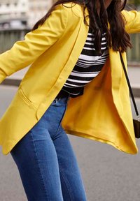 Žena se naklání dopředu, má na sobě žlutý blazer, pruhované černobílé tričko, modré džíny a nosí křížovou tašku venku.