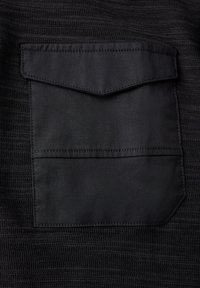 Czarna tkanina o teksturowanym wzorze, z dużą kieszenią łatana z klapą u góry, wzmocnionymi szwami i matowym wykończeniem.