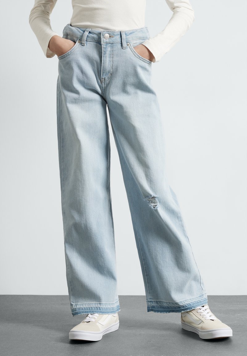 Jeans de pierna ancha en un azul claro con una cintura alta, bolsillos frontales y un detalle desgastado en la pierna izquierda. Combinados con zapatillas beiges.