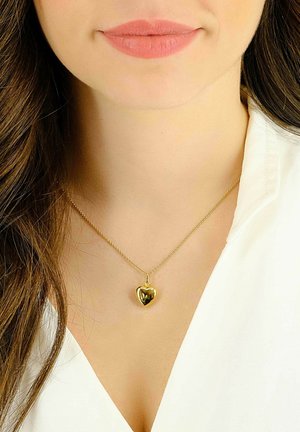 Collana con pendente a forma di cuore in oro su una delicata catena, con superficie lucida, posizionata all'altezza del collo sopra una camicia bianca.
