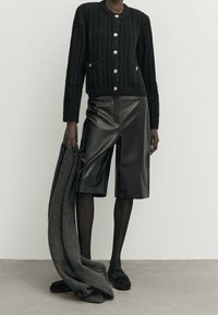 Henkilö, joka käyttää mustaa nappikardigania, mustia nahkashortseja polven korkeudelle, mustia sukkahousuja ja mustia loafereita, pitää kädessään harmaata kangasta tai takkia.