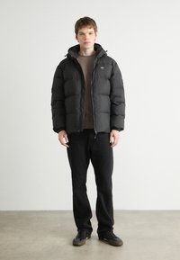 Sorth pufferjakke med hætte, lynlås lukning og elastiske manchetter, båret over en brun sweatshirt og sorte bukser. Glat, mat tekstur.