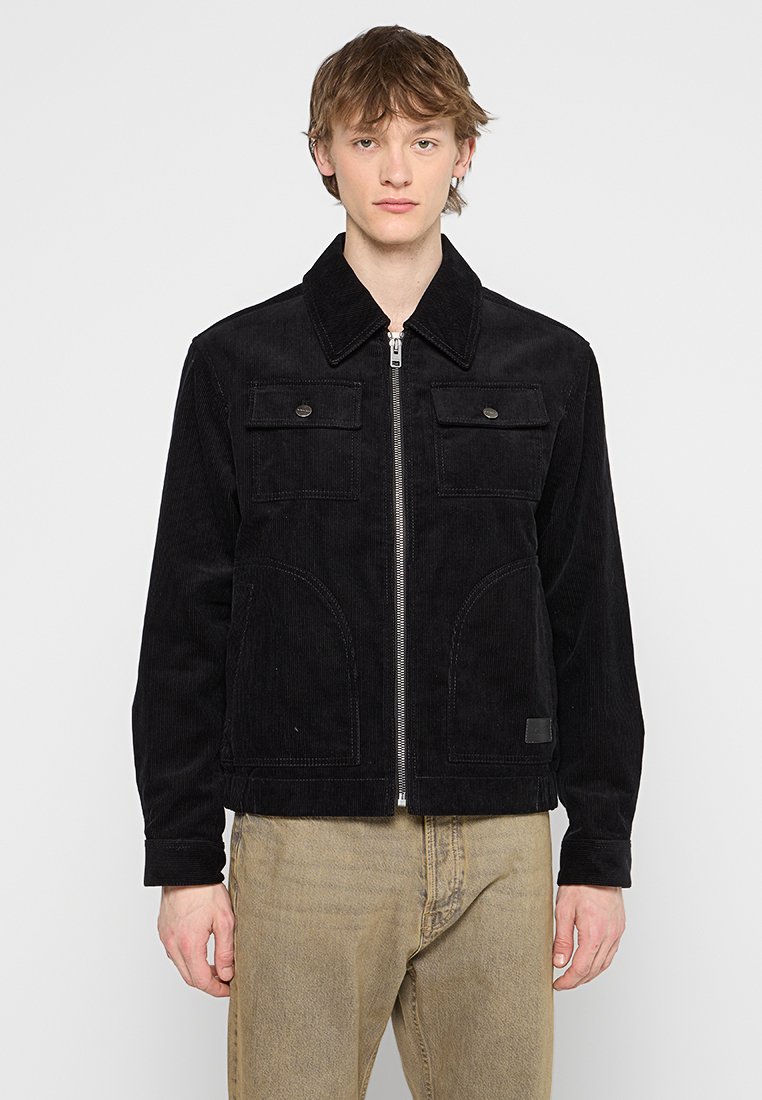 Veste en velours côtelé noir avec fermeture éclair à l'avant, grandes poches poitrine et poches latérales. Présente un col à revers et des détails de couture subtils.