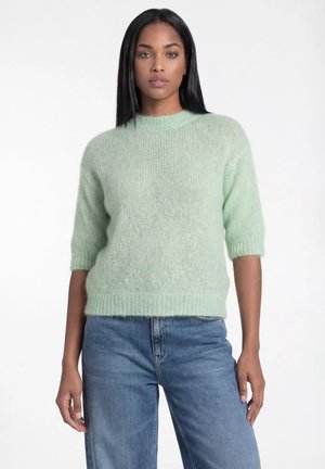 Kobieta ubrana w jasnozielony puszysty sweter z rękawami do łokci i niebieskie dżinsy z wysokim stanem na białym tle.