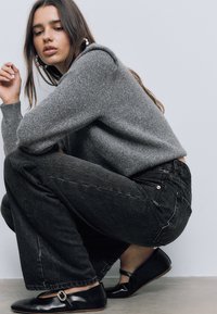 Szary sweter dzianinowy, czarne szerokie dżinsy z widocznym przeszyciem i czarne płaskie buty z detalem klamry. Model siedzi na kolanach na jasnym tle.