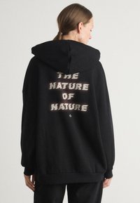 Svart hettegenser med en frontlomme i kenguru-stil, snøring i hetta og teksturert grafisk tekst "NATURSNATUR" på baksiden i kremfarge.