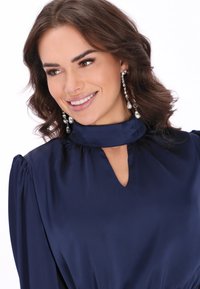 Sukienka z granatowego satynu o wysokim dekolcie i wycięciu w kształcie łezki, z bufiastymi rękawami i gładką fakturą. Widoczne są srebrne długie kolczyki.