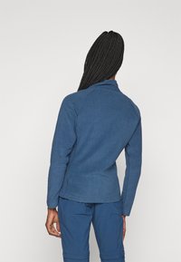 Blå fleecejacka med krage och raglanärmar, med en texturerad yta. Bärs med matchande blå byxor, vy från baksidan.