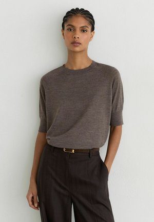 Femme aux cheveux tressés portant un haut en maille marron à manches courtes, rentré dans un pantalon marron foncé avec une ceinture fine, debout contre un mur blanc.