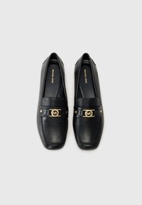 Mocassini in pelle nera con un accento hardware in oro rotondo con logo intrecciato, design piatto e superficie liscia.