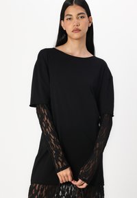 Fekete póló laza fit, kör alakú kivágással és rövid ujjakkal. Hosszú, áttetsző fekete csipkeujjak virágmintával rendelkeznek.