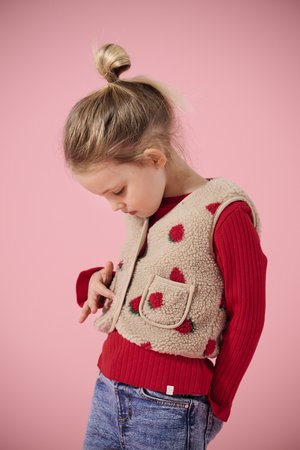 Jong meisje met blond haar in een knot, gekleed in een beige fleecevest met rode aardbeitjes over een rood truitje en een blauwe spijkerbroek, kijkt naar beneden.