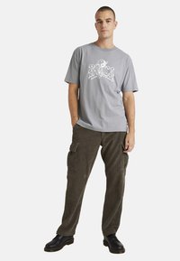 Gray cotton t-shirt featuring a white graphic print. Paired with olive corduroy cargo pants with side pockets and black shoes.